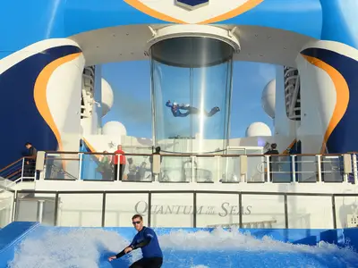 Eine Person schwebt in einem gläsernen Skydiving-Tunnel auf dem Oberdeck der Quantum of the Seas bei Tageslicht. Andere Passagiere beobachten die Szene vom Deck aus.