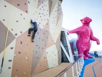 Ein Kletterer an der Kletterwand auf dem Oberdeck der Quantum of the Seas. Rechts eine große, polygonale pinke Bärenskulptur und ein Laufpfad.
