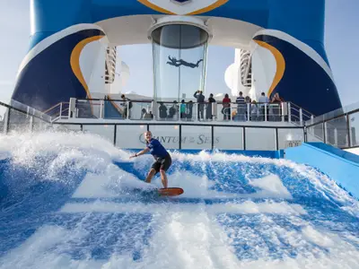 Auf der Quantum of the Seas surft eine Person auf einer künstlichen Welle. Im Hintergrund schwebt eine weitere Person in einem gläsernen Windkanal, beobachtet von vielen Zuschauern.