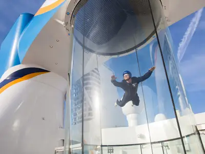 Eine Person im schwarzen Fluganzug schwebt im iFLY Windkanal der Quantum of the Seas. Der gläserne Simulator steht vor blauem Himmel mit Kondensstreifen.
