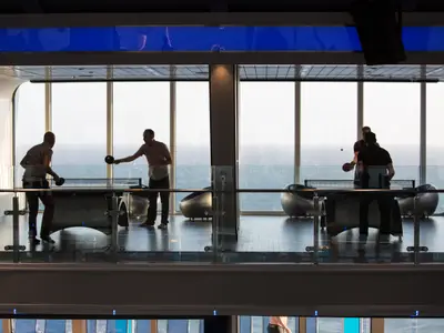 Auf der Quantum of the Seas spielen zwei Gruppen Tischtennis. Die Szene spielt in einem lichtdurchfluteten Raum mit weitem Meerblick durch große Panoramafenster. Die Spieler sind als Silhouetten erkennbar.