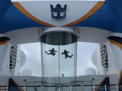 Blick auf das Deck der Quantum of the Seas mit zwei Personen im gläsernen Indoor-Skydiving-Simulator. Oberhalb des Tunnels ist das Royal Caribbean Logo zu sehen.