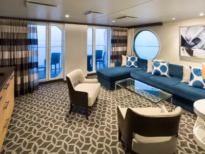 Geräumiges Wohnzimmer einer Suite der Quantum of the Seas. Blauer Sofabereich, zwei Sessel, gemusterter Teppich, sowie Fenster und runder Ausblick auf das Meer.