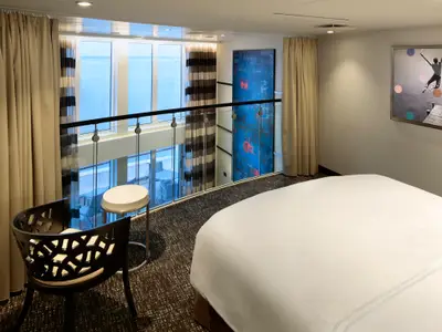 Innenansicht einer eleganten, zweistöckigen Loftkabine an Bord der Quantum of the Seas. Im Vordergrund ein weiß bezogenes Bett, dahinter eine Glasbalustrade mit Blick auf den unteren Bereich der Kabine und das offene Meer.
