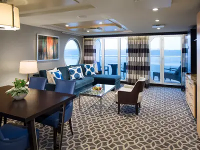 Innenansicht einer Suite auf der Quantum of the Seas. Gezeigt werden ein Essbereich mit dunklem Tisch, eine blaue Eckcouch, ein Glastisch und Fenster mit Meerblick.
