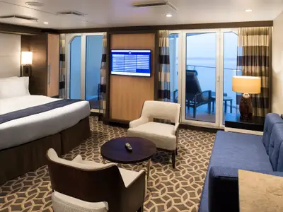 Innenansicht einer Balkonkabine auf der Quantum of the Seas: Doppelbett, Sofa, Sessel, TV. Durch die Glastür ist ein Balkon mit Meerblick und Liegestuhl sichtbar. Warme Farben.