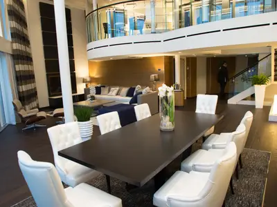Luxuriöser, zweistöckiger Ess- und Wohnbereich auf der Quantum of the Seas. Dunkler Esstisch, weiße Stühle, dahinter Sofa und Galerie. Ein Mitarbeiter steigt die Treppe.