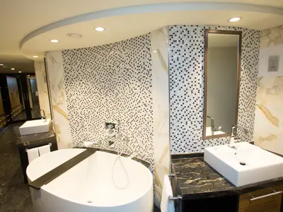 Ein elegantes Badezimmer an Bord der Quantum of the Seas mit freistehender Badewanne, zwei Waschtischen, Mosaikfliesen und einem Ankleidebereich im Hintergrund.