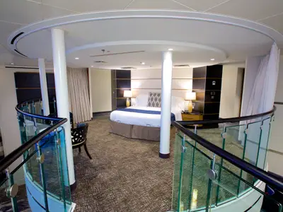 Blick von oben in einen modern gestalteten Schlafzimmerbereich der Quantum of the Seas. Ein großes Bett mit Nachttischen, Lampen und eine geschwungene Glasbrüstung sind zu sehen.