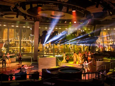 Szene einer Theateraufführung an Bord der Quantum of the Seas. Tänzer in Kostümen auf einer mehrstufigen Bühne mit opulentem Hintergrund und zahlreichen Lichteffekten. Zuschauerplätze und ein kleiner Pool sind im Bildvordergrund.