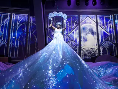 Eine Performerin in weißem, langem Kleid mit sternenübersäter Schleppe hält eine große Krone bei einer Show auf der Quantum of the Seas. Der Hintergrund zeigt blaue, stilisierte Fenster mit Mond und Sternen.