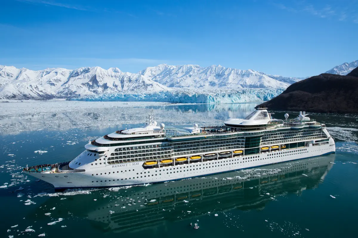 Die Radiance of the Seas mit Passagieren auf den Decks im eisigen Wasser, umgeben von schneebedeckten Bergen und einem blauen Gletscher unter klarem Himmel.