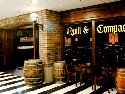 Eingang der Bar 'Quill & Compass' auf der Radiance of the Seas. Ziegelwände, Holzfässer, schwarz-weißer Boden und goldener Schriftzug.