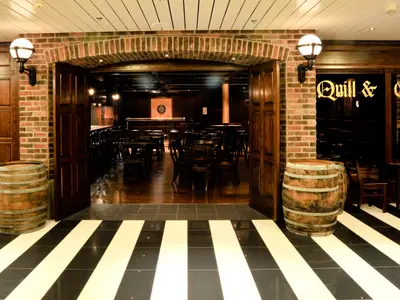 Eingangsbereich des "Quill & Compass" Pubs. Ziegelwände, Holzfässer und schwarz-weiß gestreifter Boden. Innen Tische, Stühle, Dartscheibe.