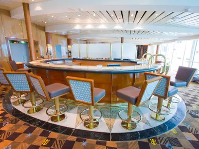 Eine leere, geschwungene Bar an Bord der Radiance of the Seas mit zahlreichen Barhockern, Holztheke und Teppichboden in einem hellen Innenbereich.