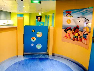 Eine bunte Kinderspielzone an Bord der Radiance of the Seas mit gelben und hellgrünen Wänden, einem blauen Boden und einer Fisher-Price Little People Grafik an der Wand. Im Hintergrund sind mehrere Türen sichtbar.