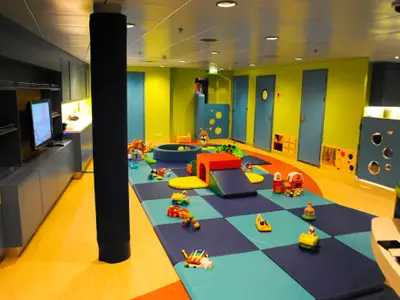 Ein bunter Spielbereich im Kids' Club der Radiance of the Seas, ausgestattet mit blauen Matten, Spielgeräten, einem Ballbad und zahlreichen Spielsachen für Kinder. Eine TV-Anlage ist links sichtbar.