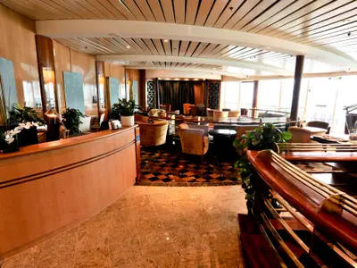 Ein heller Innenbereich auf der Radiance of the Seas zeigt eine geschwungene Holzrezeption mit Pflanzen und dahinter eine Lounge mit Polstersesseln und Tischen. Große Fenster lassen im Hintergrund viel Tageslicht herein.