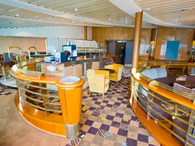 Interieur der Radiance of the Seas: Lounge-Bereich mit Selbstbedienungsbar, gelben Sesseln, Sitzecken, Holz- und Messinggeländern. Ein Mitarbeiter geht im Hintergrund.