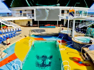Das Pooldeck der Radiance of the Seas zeigt einen großen Pool, Whirlpools, eine Videowand und Liegestühle. Die bunte Bodengestaltung sowie das Meer im Hintergrund sind deutlich zu erkennen.