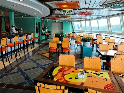 Restaurant der Radiance of the Seas: bunte Einrichtung, Bar mit Hockern, Tische mit Mosaikplatten, Panoramablick durch Fenster auf bewaldete Küste.