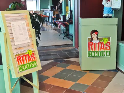 Eingang zur Ritas Cantina auf der Radiance of the Seas. Ein Menüständer und eine grüne Theke mit Logo stehen auf Fliesen. Im Hintergrund ein Restaurantbereich.