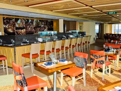 Einblick in ein Bordrestaurant der Radiance of the Seas: eine lange Theke, Esstische mit orangefarbenen Stühlen, Meerblick durch Fenster und ein Exit-Schild.