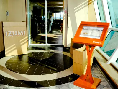 Detailansicht des Eingangs vom Izumi Restaurant auf der Radiance of the Seas. Ein oranger Menüständer und ein Holzschild stehen am gefliesten Boden. Durch die Glastür ist das Meer zu erkennen.