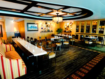 Blick in einen Speisesaal der Radiance of the Seas. Eine lange Tafel mit gepolsterter Bank, mehrere Tische mit Stühlen und Weinschränke sind zu sehen.