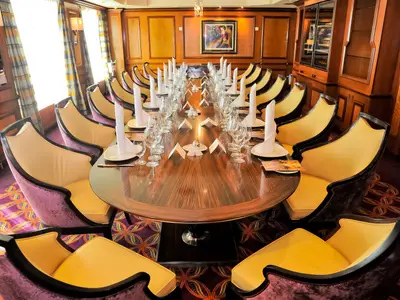 Innenansicht eines festlich gedeckten Speisezimmers auf der Radiance of the Seas. Ein langer Holztisch mit weißen Servietten, Gläsern und gelben Stühlen. Die Wände sind holzvertäfelt und mit einem Gemälde geschmückt.