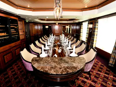 Ein eleganter, langer Speisesaal mit dunklen Holzvertäfelungen und einer großen, marmorierten Tafel. Die Tafel ist für viele Gäste mit weißen Servietten, Gläsern und Geschirr gedeckt. Seitlich sind Weinkühlschränke sichtbar. Der Raum strahlt eine luxuriöse Atmosphäre aus.