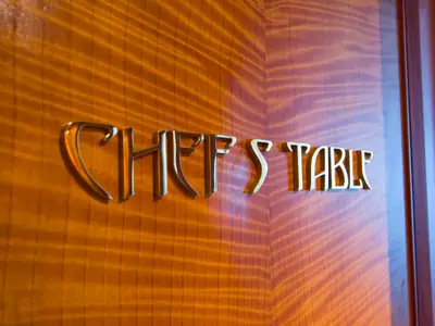 Nahaufnahme der goldenen Schrift 'CHEFS TABLE' auf einer eleganten, hellbraunen Holzwand. Indirekte Beleuchtung erzeugt weiche Schatten unter den Buchstaben.