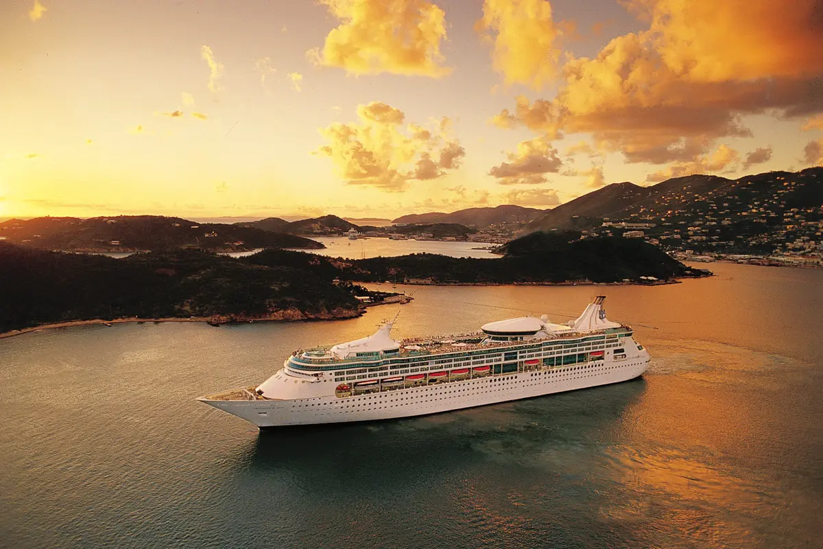 Die Rhapsody of the Seas fährt bei Sonnenuntergang durch eine tropische Bucht. Im Hintergrund sind bewaldete Inseln und eine Stadt an Hügeln zu erkennen. Der Himmel zeigt warme Gold- und Orangetöne.