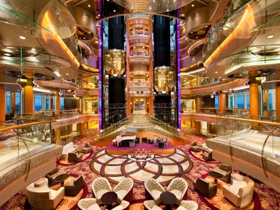 Das prunkvolle Atrium der Rhapsody of the Seas mit einer Bühne, weißem Flügel und Schlagzeug. Goldene Glaslifte und elegante Sitzbereiche auf mehreren Decks bieten Meerblick durch Fenster.