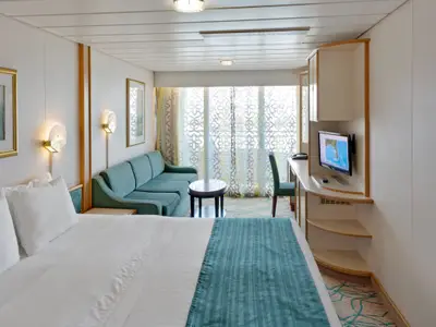 Innenansicht einer geräumigen Balkonkabine auf der Rhapsody of the Seas. Im Vordergrund ein Bett, gefolgt von einem Sofa, Schreibtisch mit TV und dem Balkonfenster. Helle Farben dominieren.