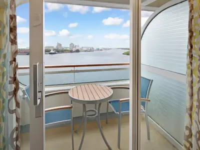 Ansicht von einer Balkonkabine der Rhapsody of the Seas mit einem Tisch und zwei Stühlen. Der Blick geht über ein Gewässer mit Industrieanlagen und einer Stadt im Hintergrund.