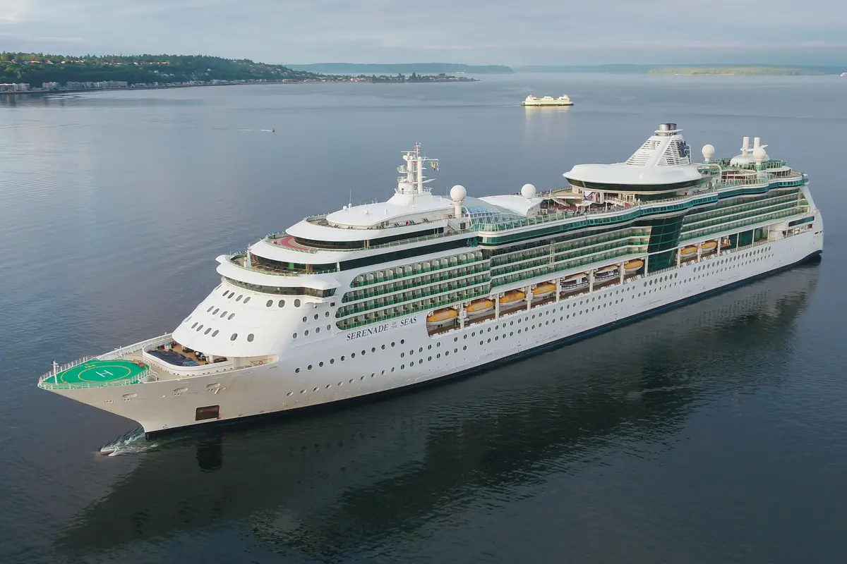 Luftaufnahme der weißen Serenade of the Seas auf ruhigem Meer. Im Hintergrund bewaldete Küste, Stadt und eine Fähre. Himmel leicht bewölkt.