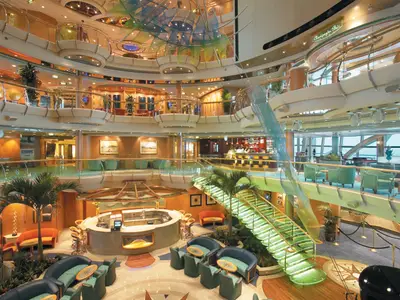 Innenansicht des mehrstöckigen Atriums der Serenade of the Seas mit Bars, grünen Sitzbereichen, einer beleuchteten Treppe und Meerblickfenstern.