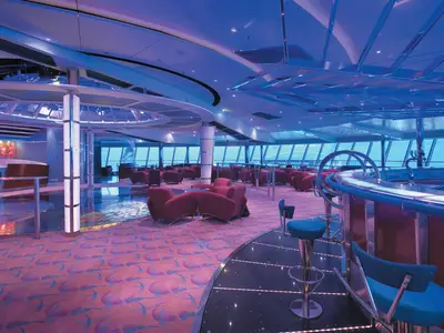 Die helle und weitläufige Lounge der Serenade of the Seas zeigt eine Bar mit Barhockern und zahlreiche rote Sessel. Große Fensterfronten bieten Meerblick, die Beleuchtung ist blauviolett.