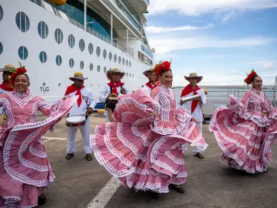 Tänzerinnen in rot-weißen Trachten und Musiker performen vor der Serenade of the Seas. Das weiße Schiff liegt am Kai bei blauem Himmel.