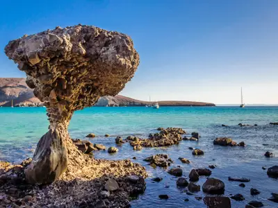 Ein pilzförmiger Felsen steht im flachen, türkisfarbenen Wasser eines Strandes. Im Hintergrund ankern mehrere Segelboote vor bewachsenen Hügeln unter klarem blauem Himmel.