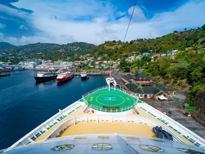Der Bug der Serenade of the Seas ist im Hafen zu sehen, mit dem grünen Helipad. Dahinter erstreckt sich eine farbenfrohe Küstenstadt an grünen Hügeln und weitere Schiffe im Wasser.