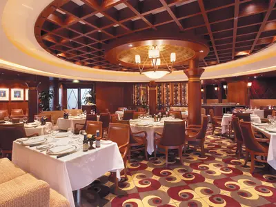 Innenansicht des eleganten Hauptrestaurants auf der Serenade of the Seas. Zu sehen sind gedeckte Tische, Ledersessel, dunkle Holzwände und eine Kassettendecke mit Kronleuchter.