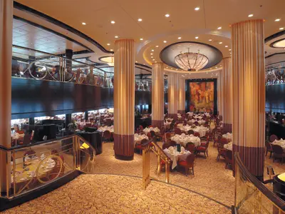 Blick in das elegante Hauptrestaurant der Serenade of the Seas. Unten viele gedeckte Tische mit roten Stühlen. Eine obere Galerie, goldene Säulen und ein großer Kronleuchter sind prägend.