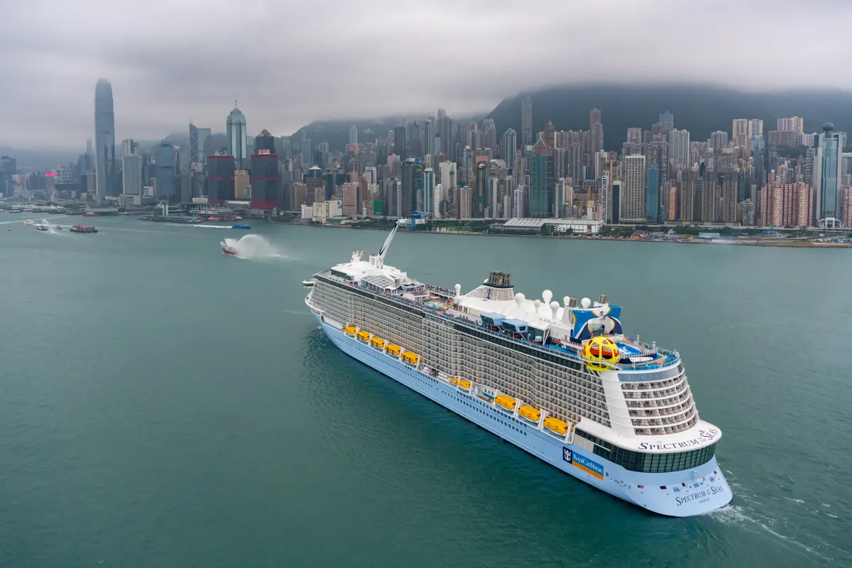 Vogelperspektive auf die Spectrum of the Seas, die in einem Hafen mit nebliger Stadtskyline und Bergen im Hintergrund schwimmt. Mehrere Boote umgeben das Schiff, eines davon spritzt Wasserfontänen.