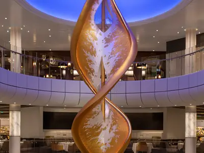Das helle Atrium der Spectrum of the Seas zeigt eine imposante, goldene Skulptur, die sich spiralförmig durch mehrere Ebenen windet. Moderne weiße Balustraden und dezente Beleuchtung prägen den Raum.
