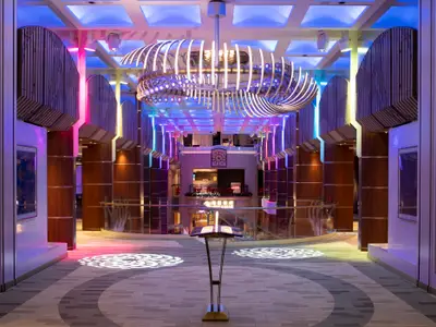 Blick in das moderne Atrium der Spectrum of the Seas. Ein großer Kronleuchter und bunte LED-Säulen prägen den Raum. Pult und Lichtmuster auf dem Boden.