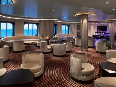 Die Lounge der Spectrum of the Seas: Sessel, Sofas, Tische und Meerblick durch Fenster. Im Hintergrund eine Bühne mit Instrumenten, teils lila beleuchtet.