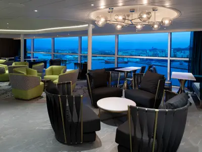 Lounge der Spectrum of the Seas. Sitzgruppen mit grünen und schwarzen Sesseln. Große Fenster blicken auf eine beleuchtete Hafenstadt bei Dämmerung. Moderner Kronleuchter und Deckenbeleuchtung.