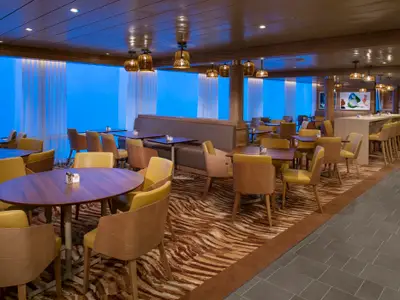 Heller Speisesaal der Spectrum of the Seas mit Tischen, Stühlen und einer Bar. Blaue Fensterfront. Holzelemente und gemusterter Teppich.
